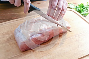 Rind from a raw roast pork is incised in a diamond shape with a sharp knife on a wooden kitchen board, preparation for cooking a