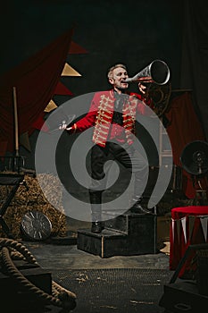 Ringmaster, magician in red coat shouting into megaphone over dark retro circus backstage background. Announcement of