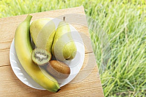 Ripe banana, kiwi fruit and pear on wooden table on background of green grasses. Rustic lifestyle concept. Top view. Copy space