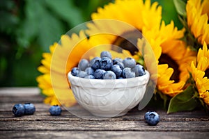 Ripe blueberries in white bowl with sunflower bouquet on wooden table, summer theme