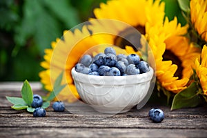 Ripe blueberries in white bowl with sunflower bouquet on wooden table, summer theme