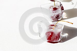 Ripe cherries in ice cubes on white background with hard sunlight, dark shadow, glare on glass. Preservation of vitamins for