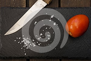 A ripe cherry stupendous tomato on a black slate along with salt flakes and a wooden handled kitchen knife