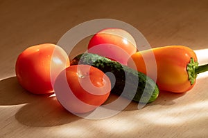 Ripe cucumber, juicy tomatoes and sweet peppers on a light wooden table in the sun. Close up