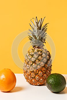 Ripe fresh pineapple, orange and avocado fruit on white table isolated on bright yellow wall background in studio