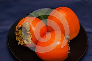 Ripe golden three persimmons on plate, dark background. These sweet and pulpy fruits can be eaten fresh, dried, raw, or