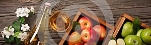 Ripe green and red apples in wooden box with branch of white flowers, glass and bottle of cider on a wooden table