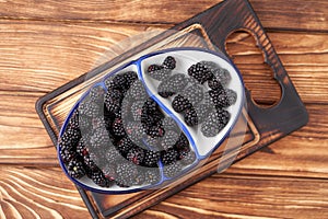 Ripe, juicy large organic blackberry in a white dish with a blue border on a cutting board on a wooden background. Vegan food