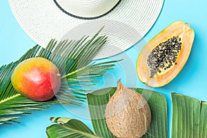 Ripe Juicy Mango Halved Papaya Coconut on Large Palm Leaf Straw Sun Hat on Blue Background. Summer Vacation Relaxation Fashion