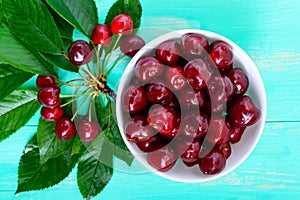 Ripe juicy red cherries in a ceramic bowl on a bright wooden background, among the leaves.