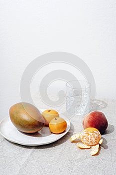 Ripe mango on a plate, tangerines, peach fruit and a glass of water on a table against white wall. Still life