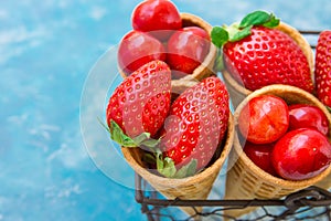 Ripe organic strawberries, glossy sweet cherries in waffle ice cream cones in wire basket, light blue background