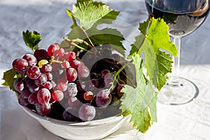 Ripe pink grapes in a white bowl and one wineglass with a red wine on white table. Selective focus