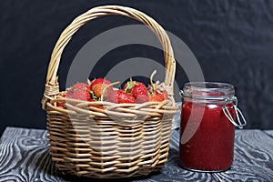 Ripe red strawberries in a wicker basket. Nearby is an open can of strawberry jam. Against the background of pine boards painted