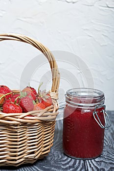 Ripe red strawberries in a wicker basket. Nearby is an open can of strawberry jam. Against the background of pine boards painted