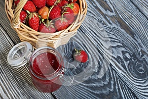 Ripe red strawberries in a wicker basket. Nearby is an open can of strawberry jam. Against the background of pine boards painted