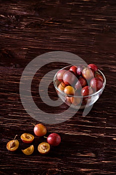 Ripe sweet plum fruits with water drops in glass bowl near to scattered sliced half plums on dark moody wood table background