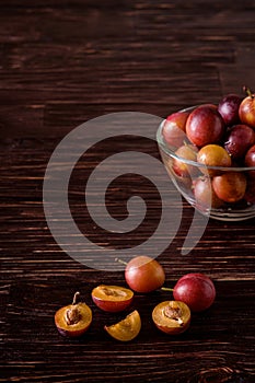 Ripe sweet plum fruits with water drops in glass bowl near to scattered sliced half plums on dark moody wood table background