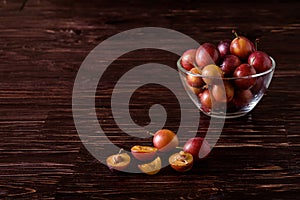 Ripe sweet plum fruits with water drops in glass bowl near to scattered sliced half plums on dark moody wood table background