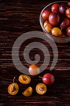 Ripe sweet plum fruits with water drops in glass bowl near to scattered sliced half plums on dark moody wood table background