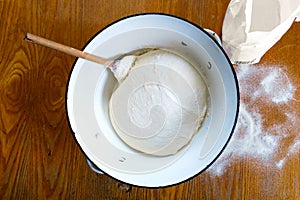 Rising yeast dough with wooden spoon in white enamel metal bowl