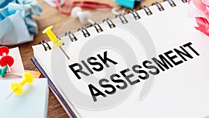 RISK ASSESSMENT - text on a notepad with wrinkled paper and paper needles on wooden background