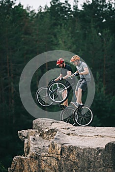 risky trial bikers standing on back wheels on rocky cliff with blurred pine forest