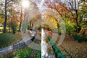 River and bicycle path in the city park in autumn. Sun rays shining through the branches of trees.