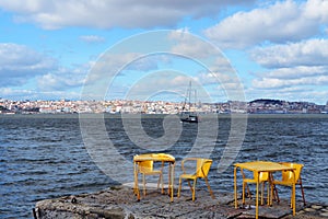 River boat on the Lisbon background. Yellow chairs and tables in riverside cafe in Almada. Portugal.