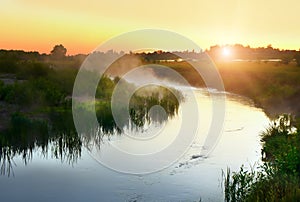 River in the early morning at dawn. Delicate dawn sky and fog rising above the water, lush greenery on the banks.