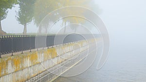 River embankment with a fence and autumn trees in fog.