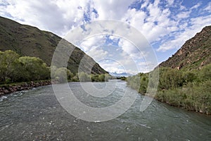 River flowing between green hills with snow-capped mountain peaks on the horizon against a cloudy sky