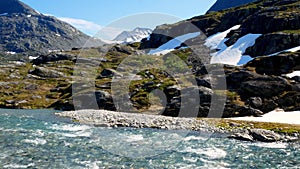 River flowing rapidly from snow melting on the high mountain rocky terrain in Norway on a sunny day. Snow topped peaks are seen.