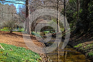 A river flowing under a brown wooden bridge in the garden surrounded by lush green grass, trees and plants with bare winter trees