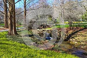 A river flowing under a brown wooden bridge in the garden surrounded by lush green grass, trees and plants with bare winter trees