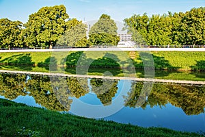 Mestom preteká rieka. Na pokojnej hladine sa odrážajú brehy rieky. Slovenská rieka Nitra za jasného slnečného dňa