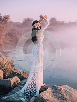 A river nymph in a white lace dress stands on a rock by the lake. The princess has a beautiful wreath with seashells