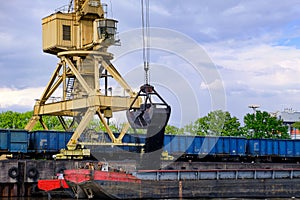 River port crane with clamshell or griper loading coal to river drag boats or barges moored by pier on cloudy day