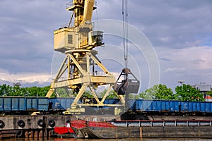 River port crane with clamshell or griper loading coal to river drag boats or barges moored by pier on cloudy day