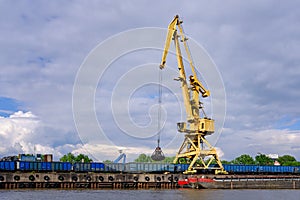 River port crane with clamshell or griper loading coal to river drag boats or barges moored by pier on cloudy day