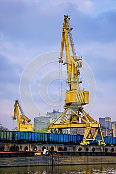 River port crane loading open-top gondola cars on cloudy day. Empty river drag boats or barges moored by pier, Empty