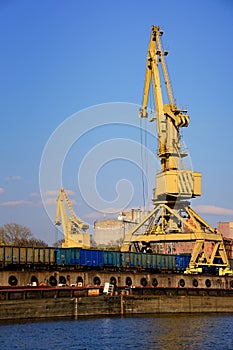 River port crane loading open-top gondola cars on sunny day. Empty river drag boats or barges moored by pier, Empty cars