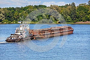 A river tugboat with an empty rusty barge goes crosses a wide river