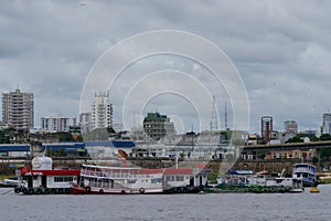 River view of a ferry refueling at a floating fuel station at the port of Manaus, with city skyscrapers visible in the background