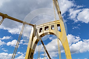 Riveted metal self anchored suspension bridge painted yellow against a bright blue sky with white puffy clouds