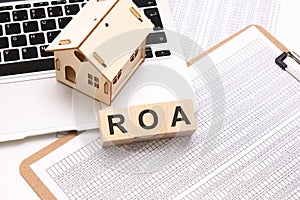 ROA - Return On Assets word on wooden cubes on a white background with a table, laptop keyboard and a small wooden house