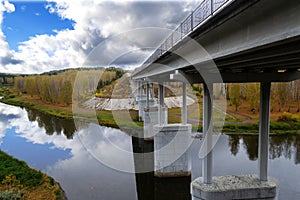 Road bridge over the river with supports on the background of the autumn forest and colorful sky with clouds.