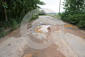 The road is full of potholes and waterlogged. The old asphalt road surface is rough and bumpy and in need of repair.