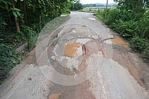 The road is full of potholes and waterlogged. The old asphalt road surface is rough and bumpy and in need of repair.