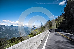 Road on the hill side running through forest with mountains, blue sky, and clouds.